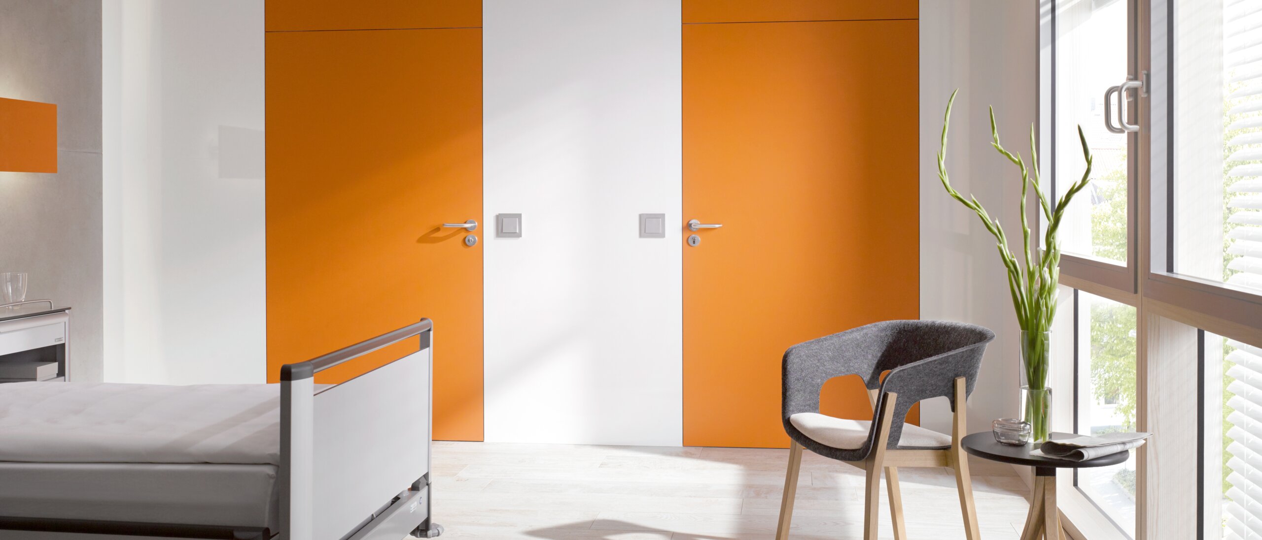 White hospital room with two orange doors fitted with a lever handle in the color grey made of polyamide